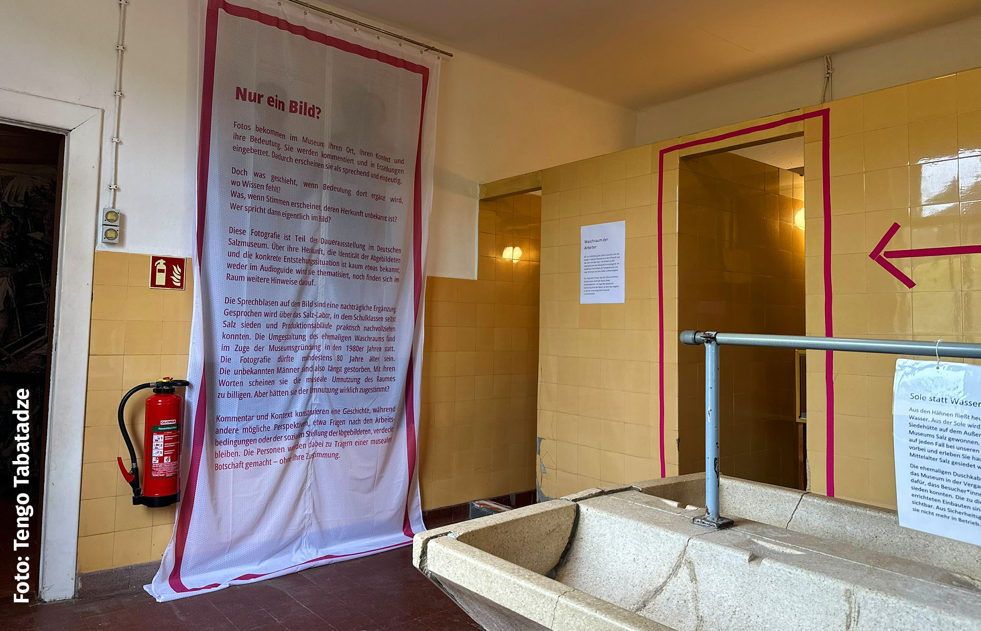 Blick in den Waschraum, der mit mehreren Interventionen neue Themen anspricht: ein Duschvorhang mit Text verdeckt das Bild zweier Männer im Waschraum, pinke Pfeile und Rahmen weisen auf die Intervention hin.